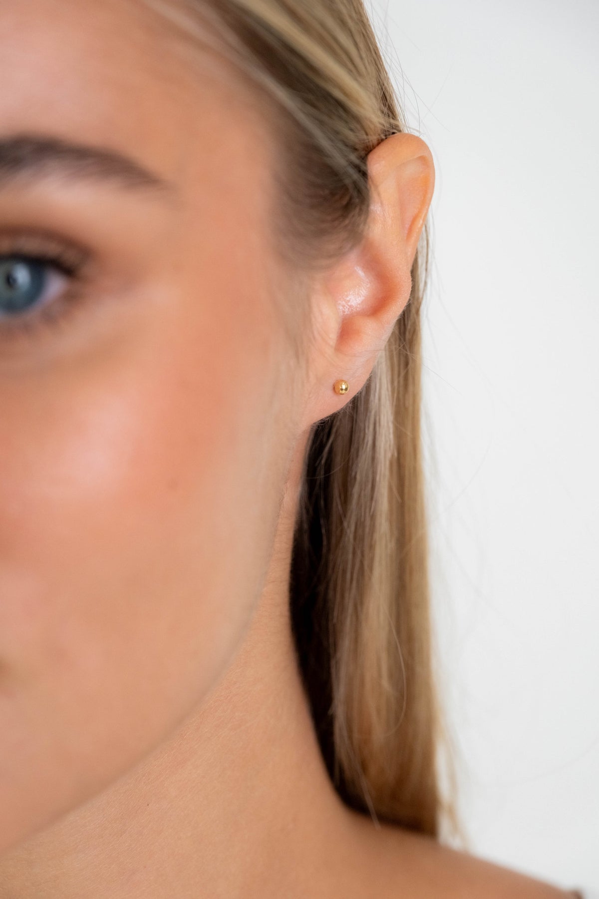 Close-up of a woman's ear wearing a tiny gold stud earring against a plain light background.