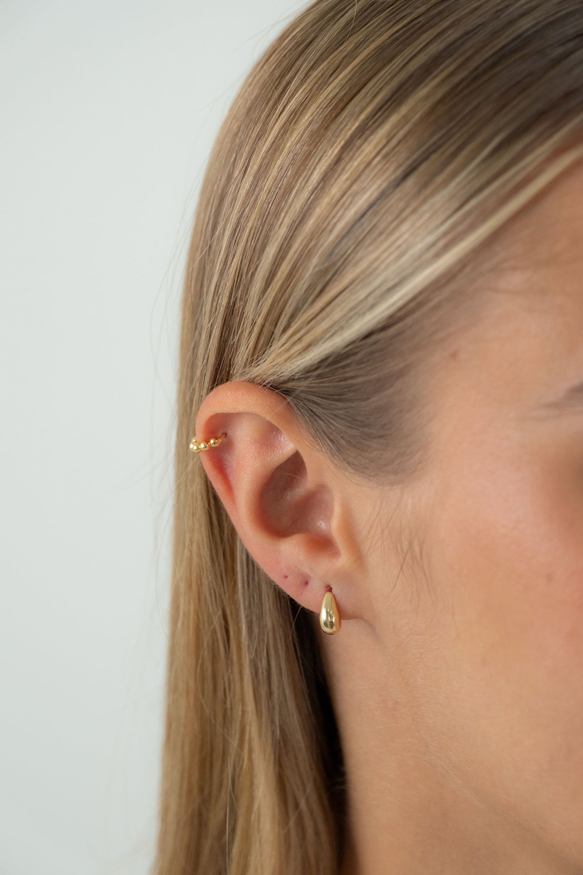 Close-up of a woman's ear wearing a small gold hoop piercing on the helix and a tiny gold teardrop stud earring, against a plain light background.