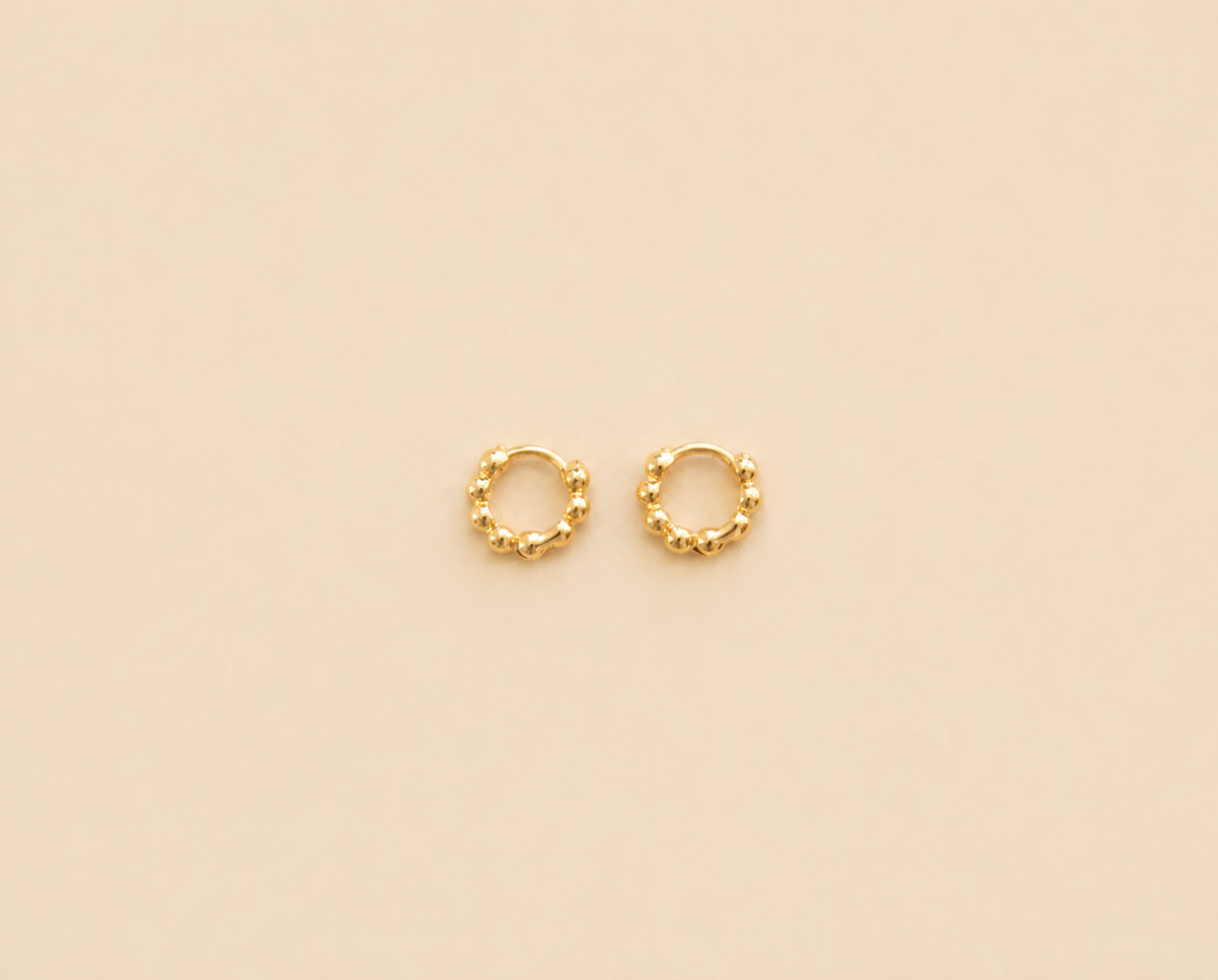 A pair of small gold huggie hoop earrings with a textured, beaded outer edge, displayed on a solid beige background.